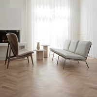 Contemporary grey sofa with a minimalist brown accent chair on a warm herringbone wood floor in a bright room.