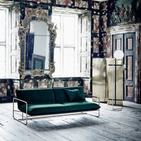 Emerald green velvet sofa against a vintage floral wall, featuring a sleek silver frame and blue accent pillow.