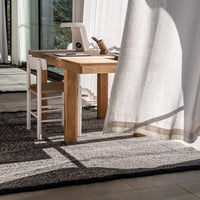 Light wood desk and white chair on textured black and white rug, complemented by sheer flowing curtain.