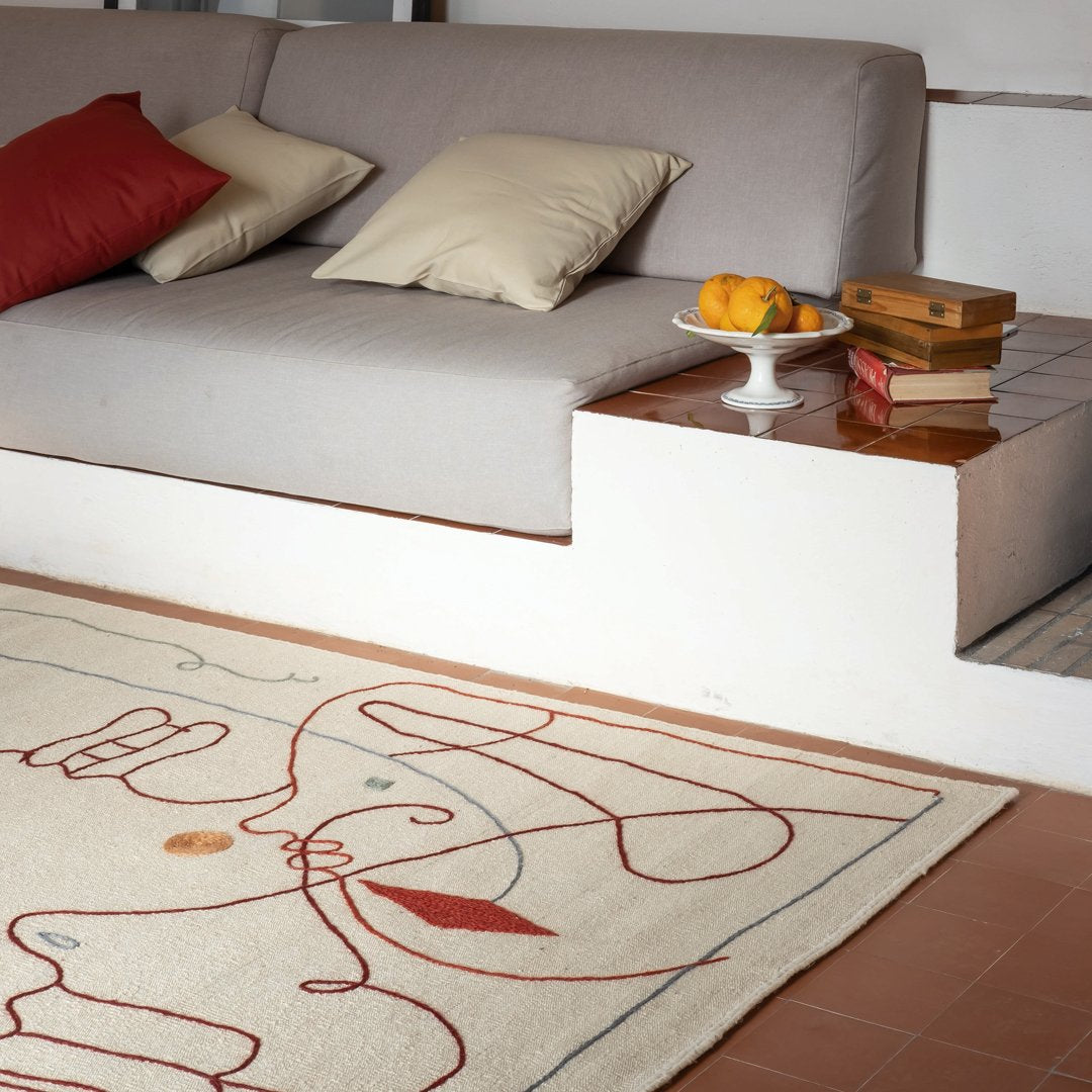 Neutral beige rug with abstract red line patterns, complemented by warm earth tones from sofa pillows and wooden decor.