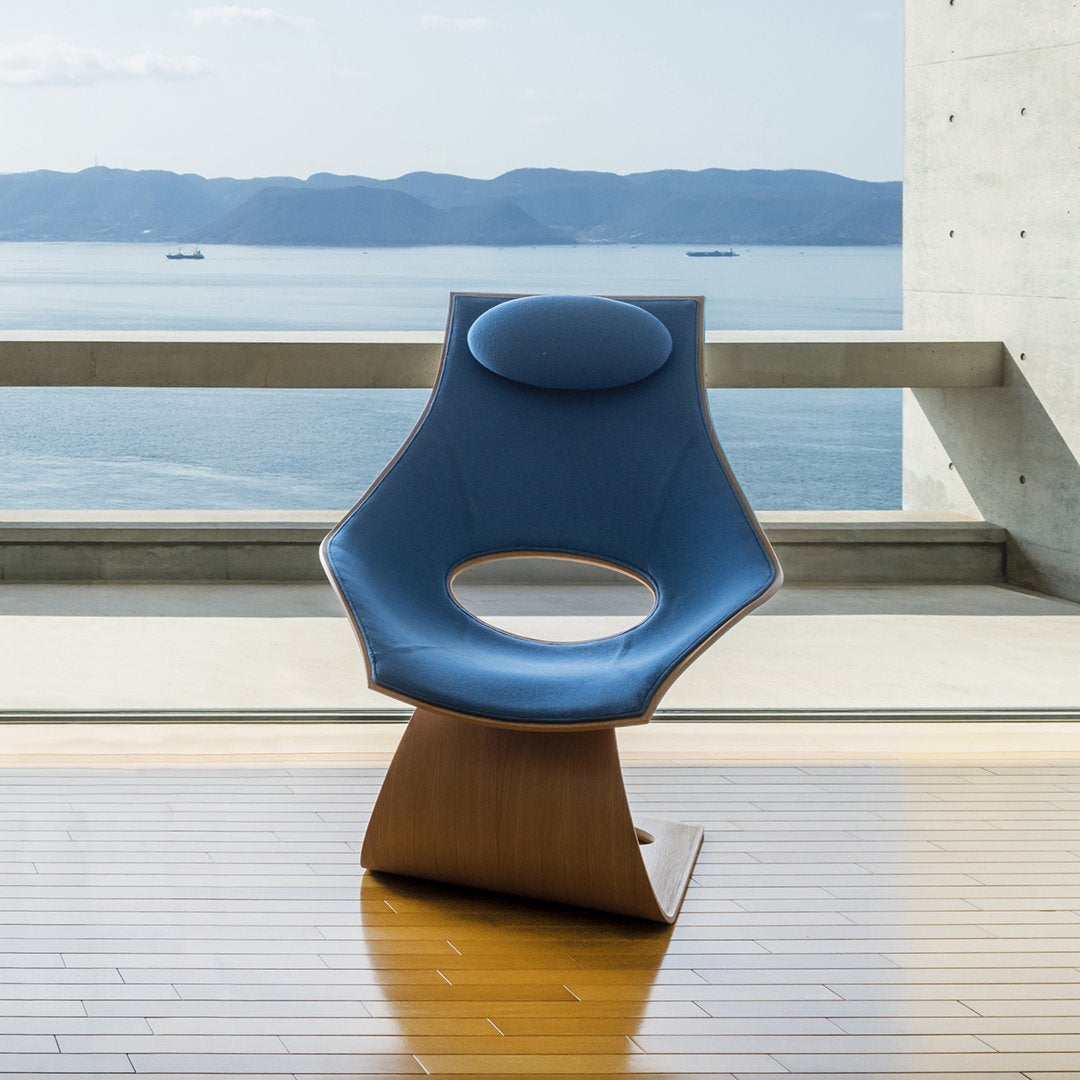 Blue ergonomic chair with a curved design and a smooth wooden base, set against a serene coastal backdrop.
