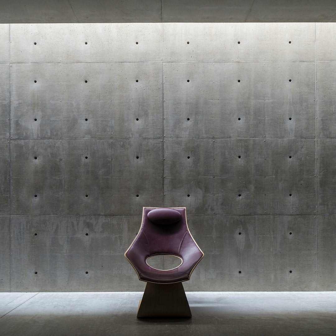 Modern chair in soft purple with a minimalist design, set against a textured concrete wall and dim lighting.