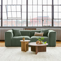 Green textured sofa with light gray and brown accent pillows, paired with round wooden coffee tables. Modern, cozy decor.