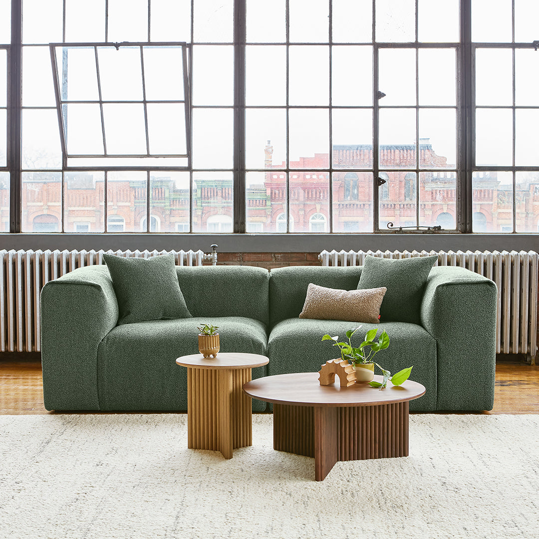 Green textured sofa with neutral accent pillows, paired with wooden side tables in light and dark brown hues.