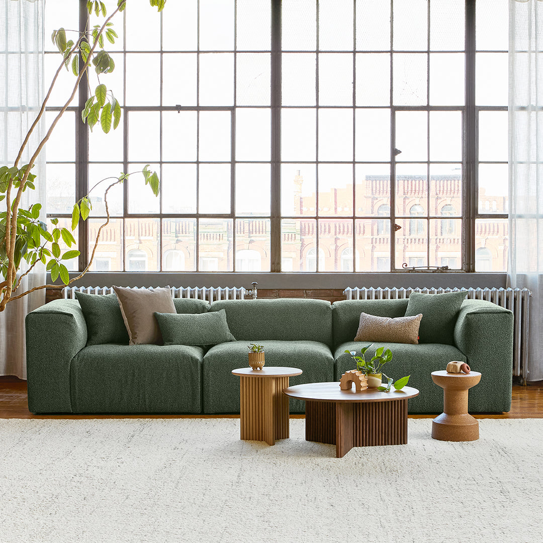 Green textured sofa with beige and brown accent pillows, paired with wooden coffee tables in a bright, airy room.