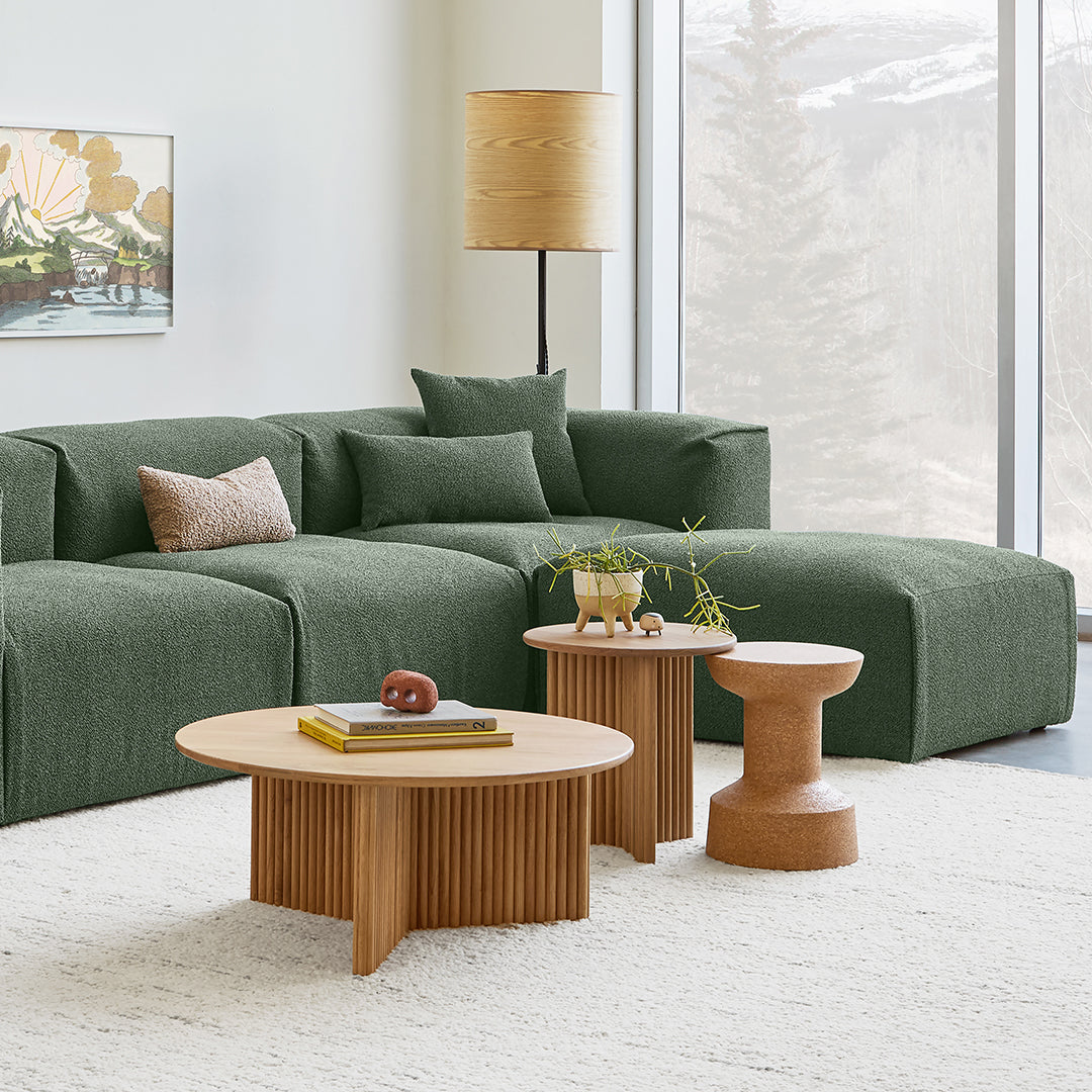 Green textured sectional sofa with soft beige pillows, paired with light wood and round table designs on a plush rug.