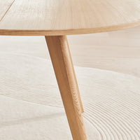 Light wooden round table with smooth finish and minimalistic design on a soft, textured light beige rug.