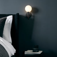 Modern wall sconce with a round white glass shade on a sleek black mount, against a dark gray wall.