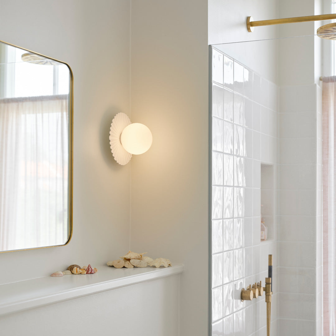 Modern wall sconce with a round white glass shade, framed by a textured, scalloped white backplate against a light bathroom backdrop.
