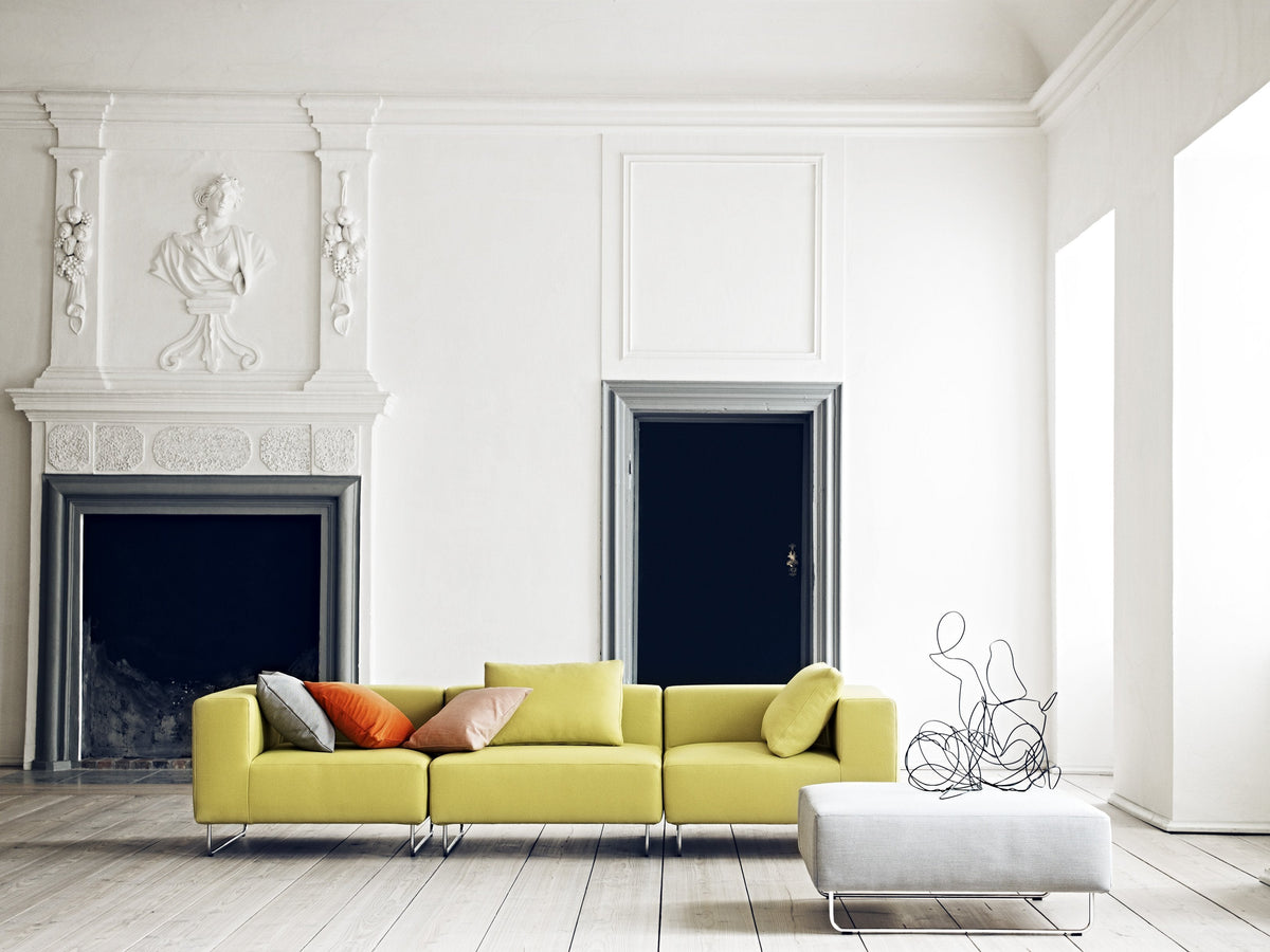 Bright yellow sectional sofa with colorful pillows, set against a minimalist white interior and light wooden floor.