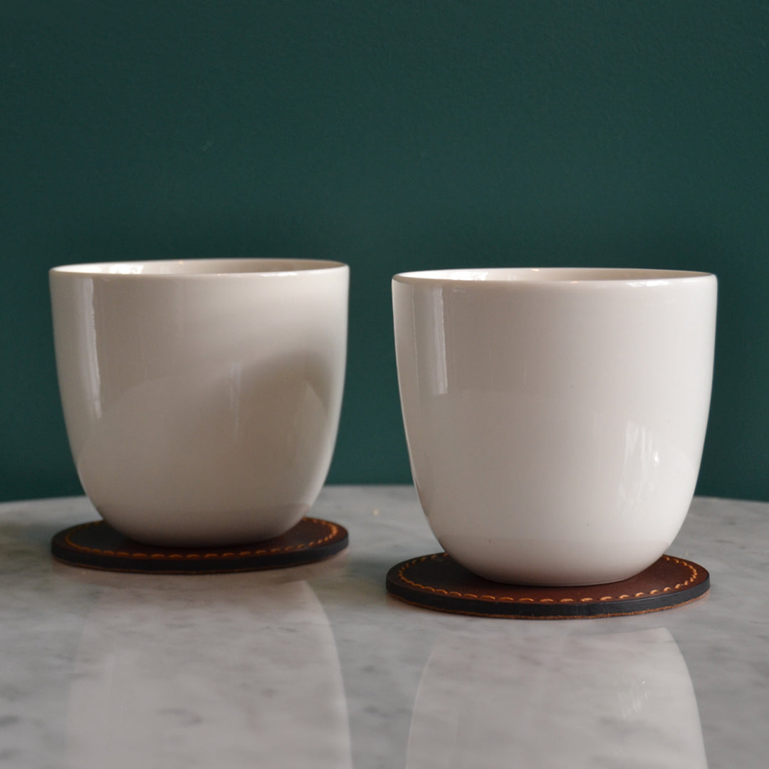 Glossy white ceramic mugs on brown leather coasters against a green wall, creating a modern and elegant contrast.