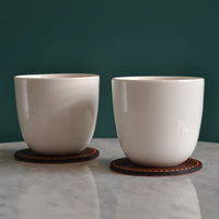 Glossy white ceramic mugs on brown leather coasters against a green wall, creating a modern and elegant contrast.
