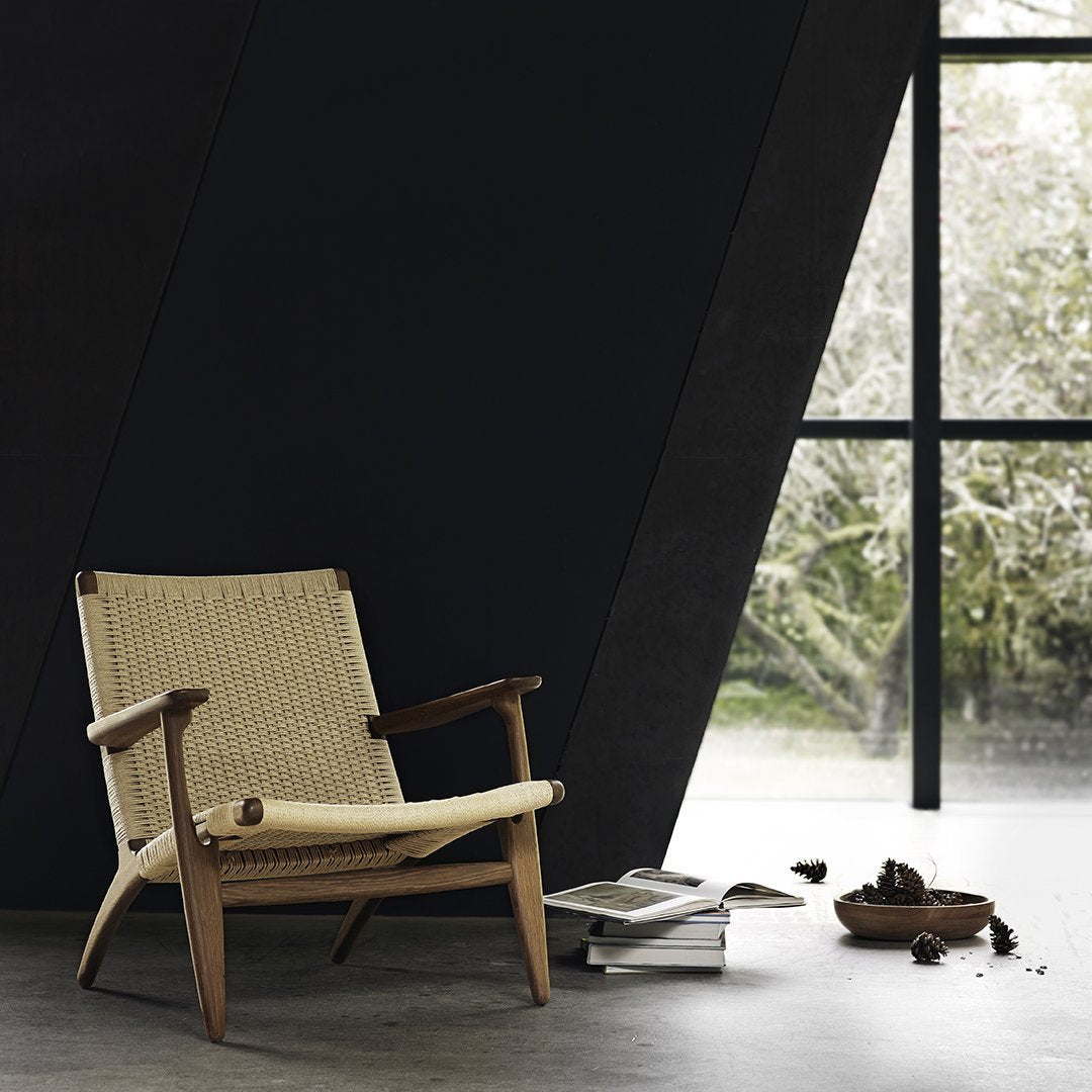 Beige woven fabric lounge chair with light wood frame, set against a dark geometric wall and natural greenery outside.