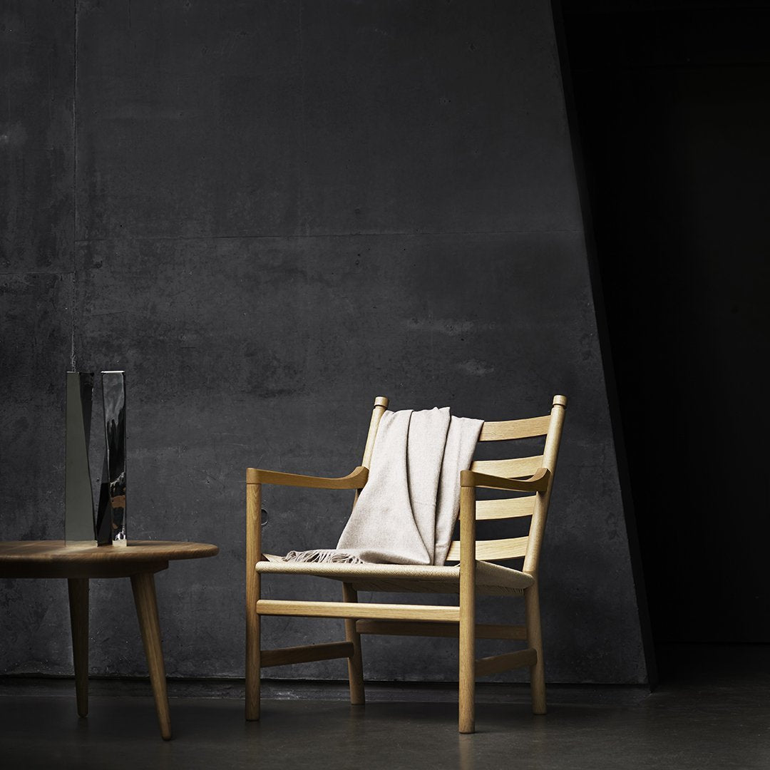 Natural wood chair with a light cream blanket, set against a dark textured background. Minimalist design highlights.