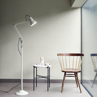 White floor lamp beside a wooden chair and black side table against a soft green wall and large window.