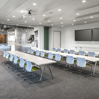 Modern conference room featuring light blue chairs, white tables, and gray carpet with green accents. Brightly lit space.