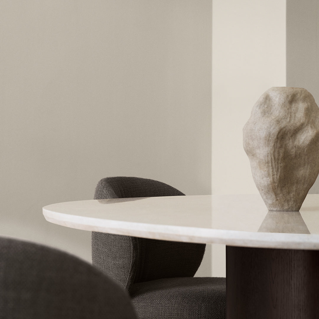 Cream round table with smooth surface, surrounded by dark gray upholstered chairs and a textured stone vase.