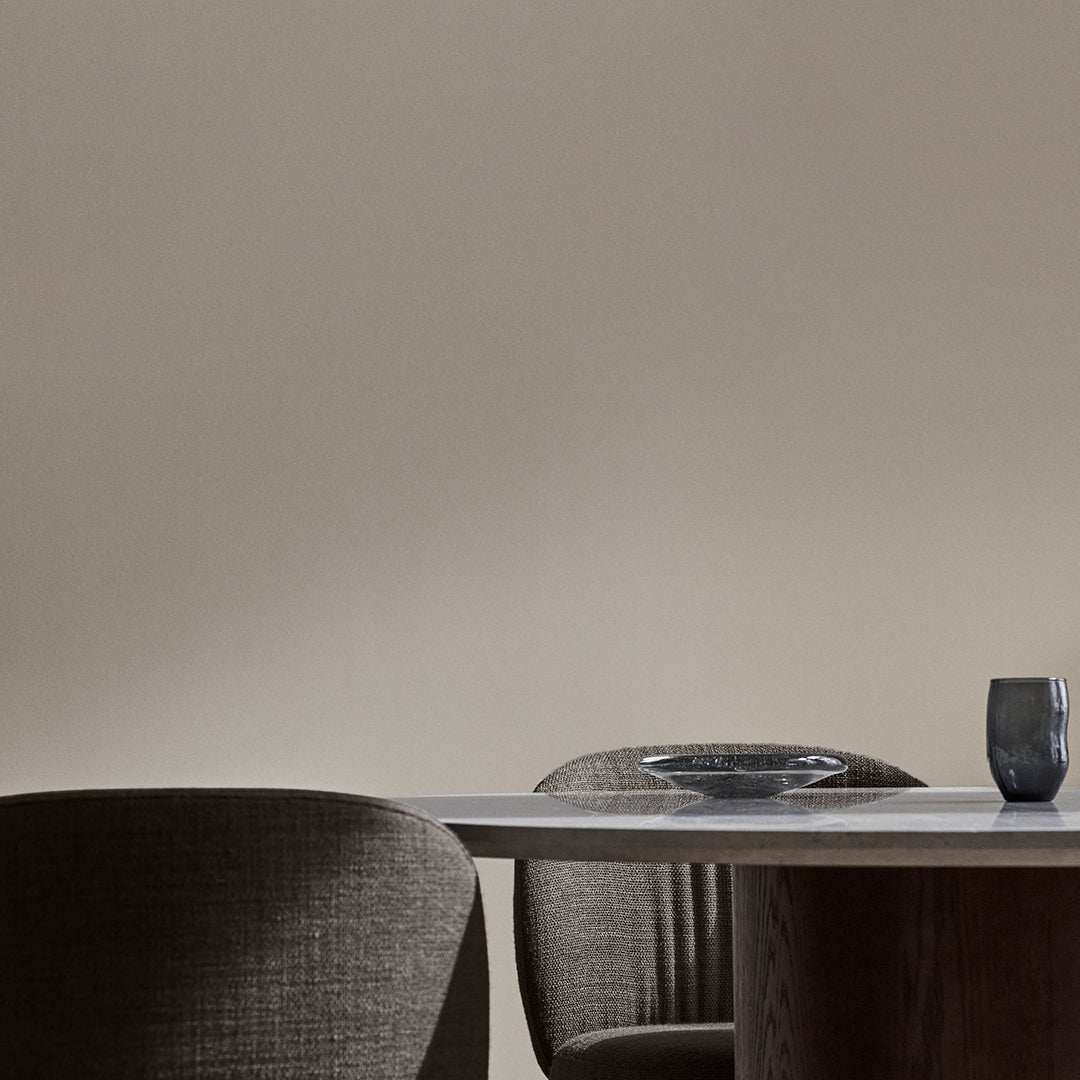 Neutral beige wall with dark brown textured chairs and a sleek wooden table, accented by a glass plate and dark tumbler.