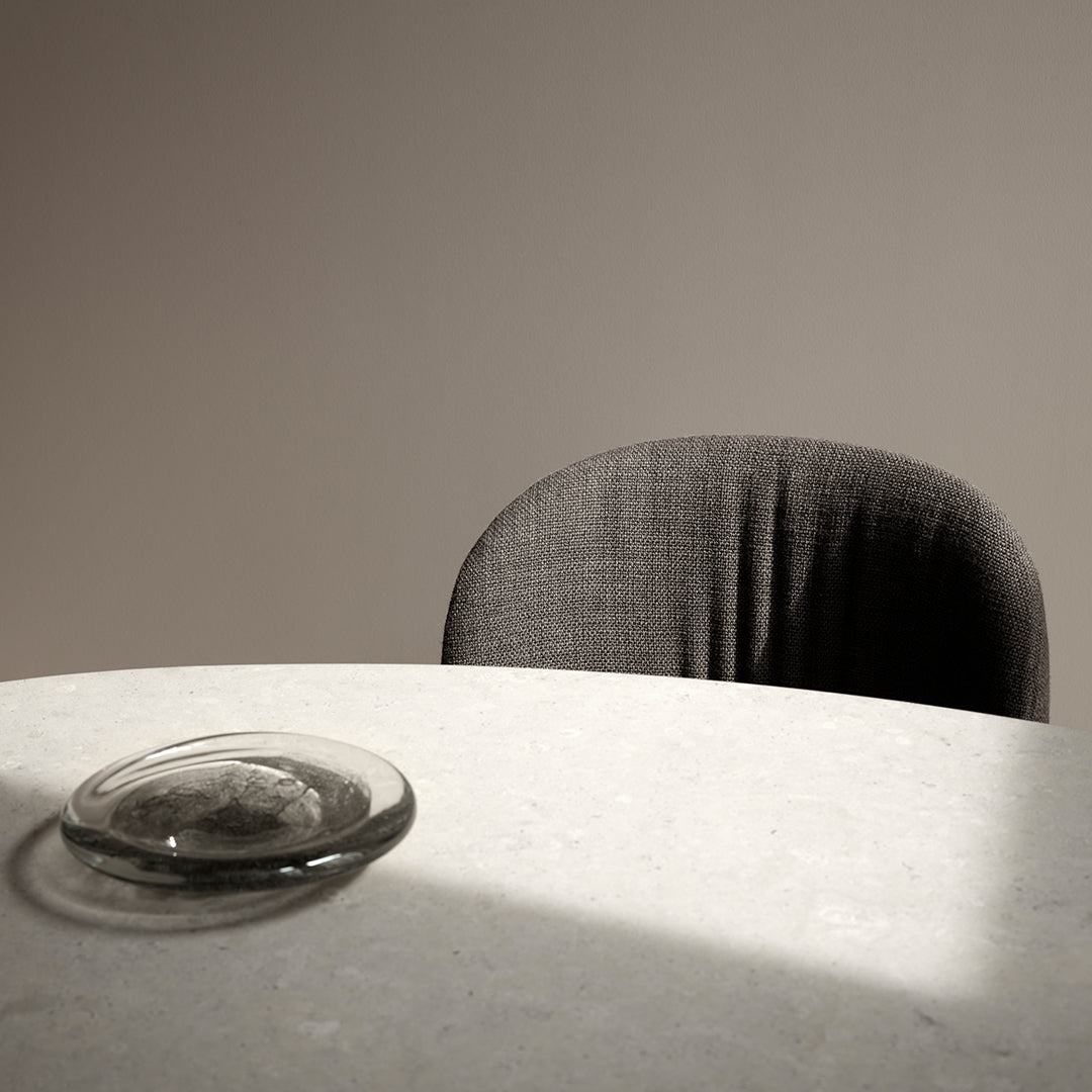 Gray textured chair against a soft beige wall, with a clear glass dish on a light stone table.