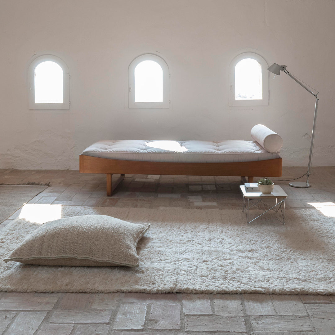 Light wood chaise lounge on a soft, textured white rug, with subtle beige and gray pillow accents.