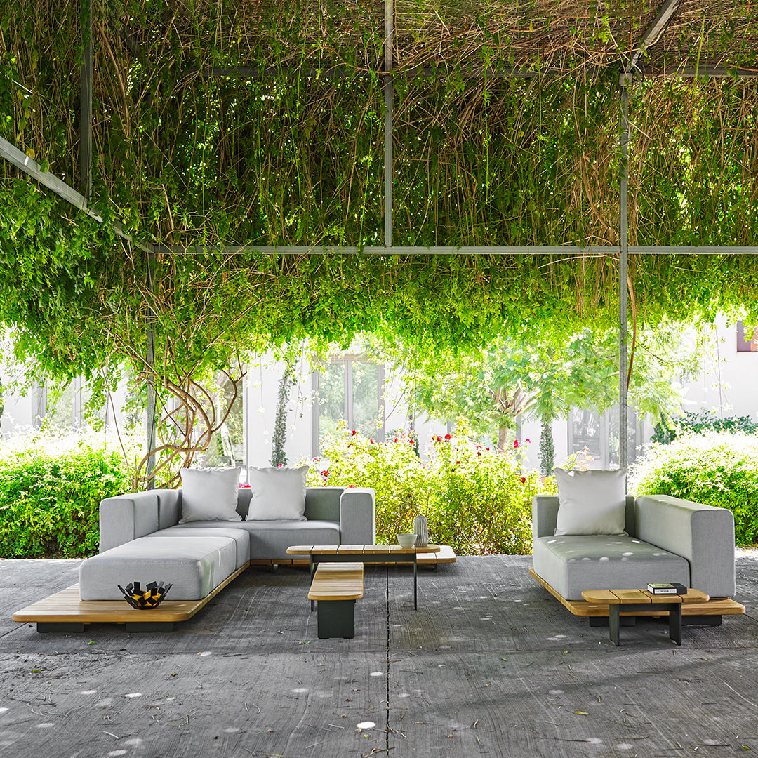 Modern outdoor sectional sofa in gray with white cushions and natural wood accents, set against lush green foliage.