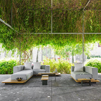 Modern outdoor sectional sofa in gray with white cushions and natural wood accents, set against lush green foliage.