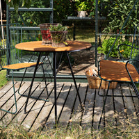 Round wooden table with black metal legs, paired with matching chairs, set against a lush garden backdrop.