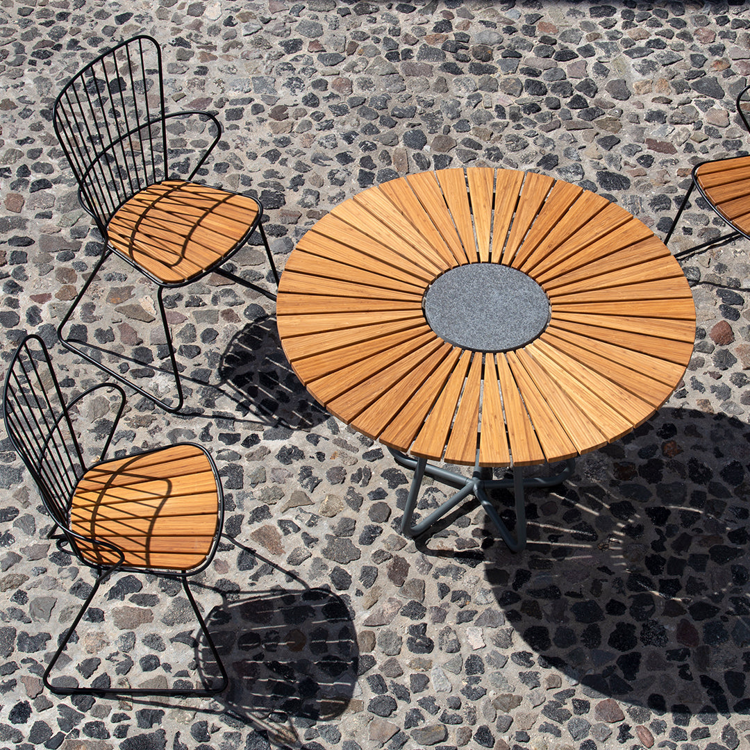 Round wooden table with a gray center, surrounded by black wire chairs with warm wooden seats, on a cobblestone surface.