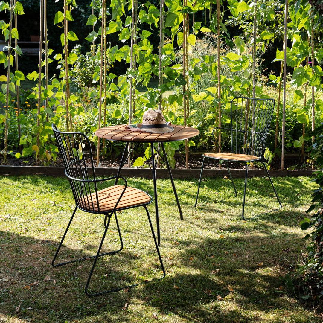 Rustic wooden table with black metal wire chairs in a vibrant green garden setting. Natural colors and textures.
