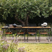 Black metal dining table with wooden accents, surrounded by patterned wire chairs in a lush green garden setting.