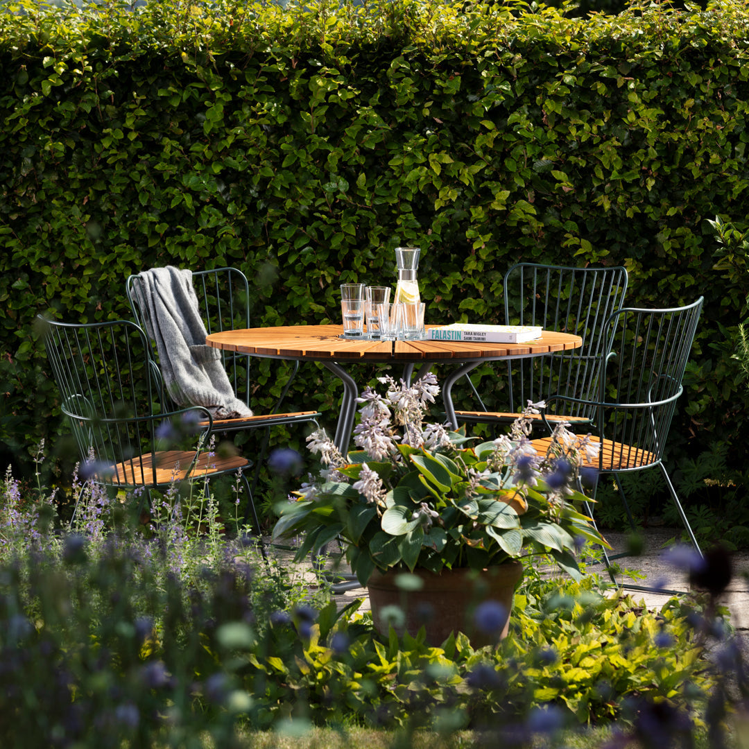 "Outdoor table set with a wooden top, green wire chairs, and vibrant plants, surrounded by lush greenery."
