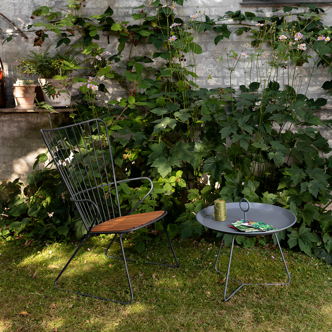 Black wire chair with wood seat, paired with a round gray table, surrounded by lush green foliage.