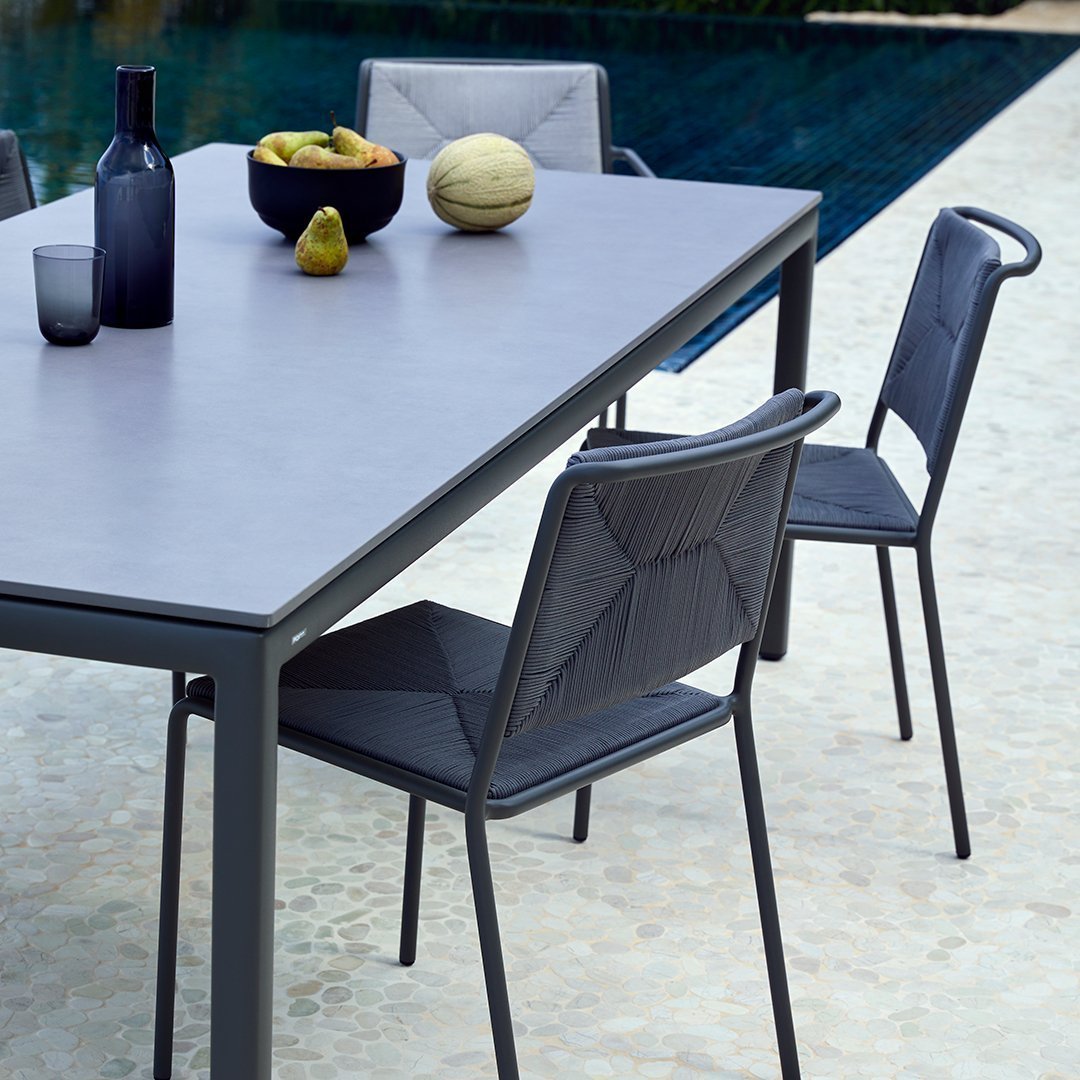 Gray outdoor dining table with black patterned chairs, surrounded by fruits and glassware near a pool.