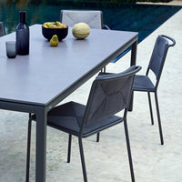Gray outdoor dining table with black patterned chairs, surrounded by fruits and glassware near a pool.
