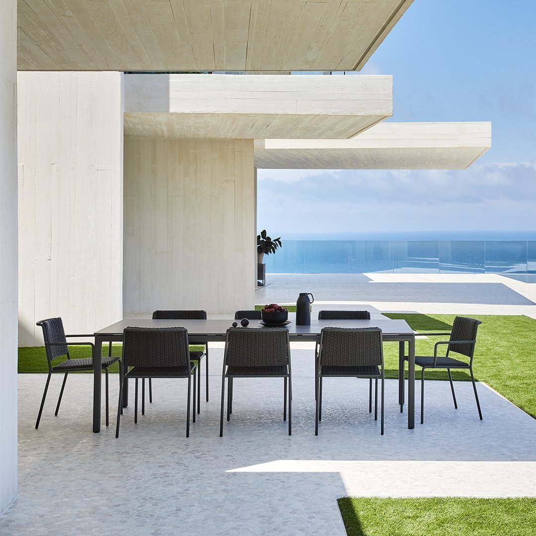 Modern outdoor dining set with sleek black chairs and a gray table, set against a backdrop of greenery and blue ocean.