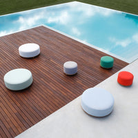 Colorful round poufs in mint, grey, white, green, and red arranged on a wooden deck by a pool. Modern design.