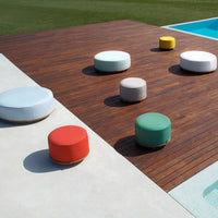Colorful outdoor poufs in pastel shades of blue, gray, green, yellow, and red arranged on a wooden deck by a pool.