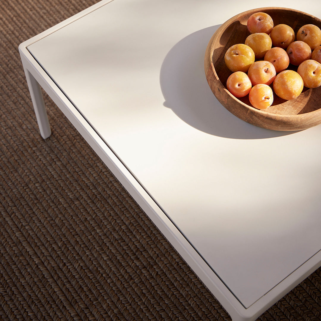 White modern coffee table with a sleek design, featuring a round wooden bowl filled with yellow apples on a textured brown rug.