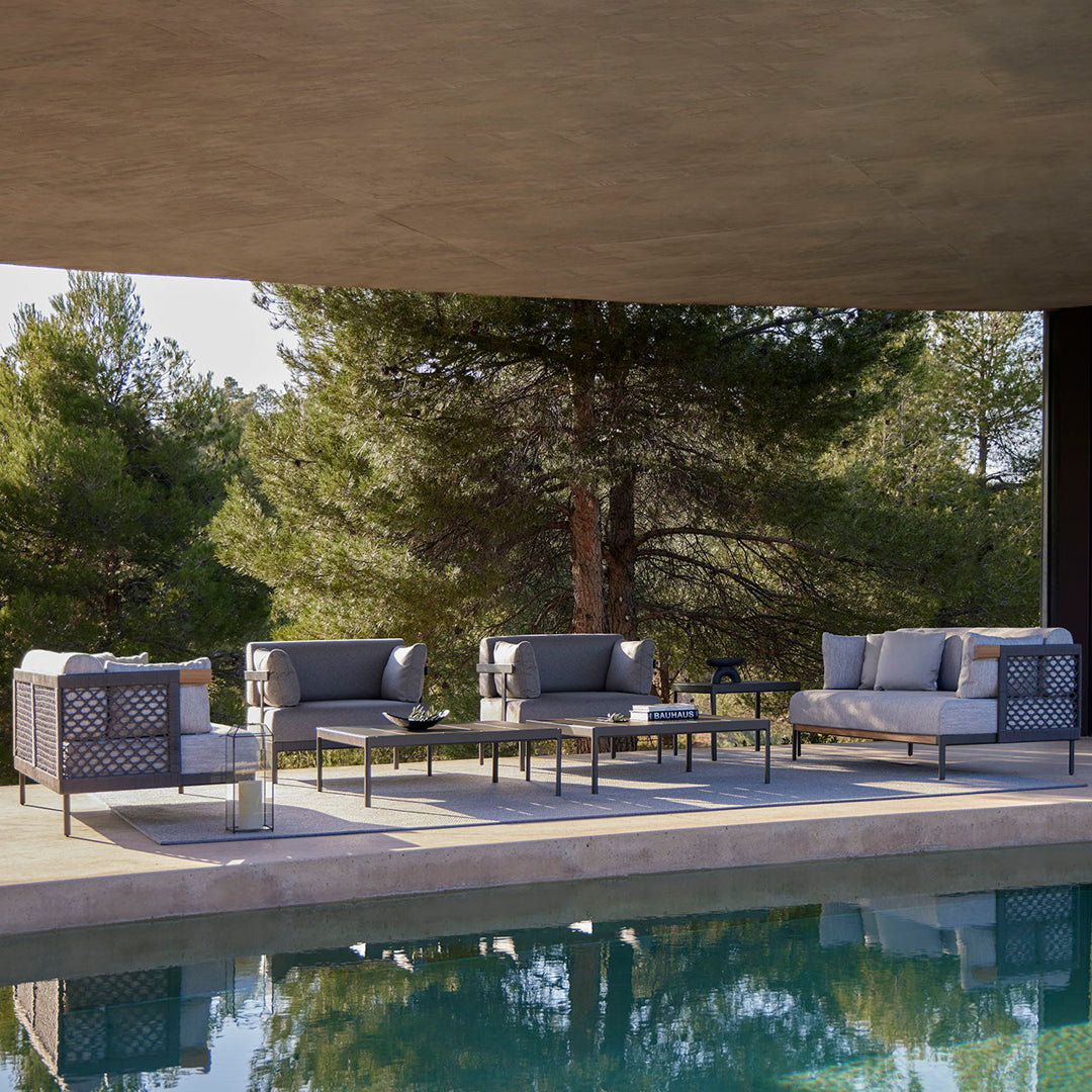 Sleek gray patio furniture with subtle geometric patterns, surrounded by green trees and a blue pool.