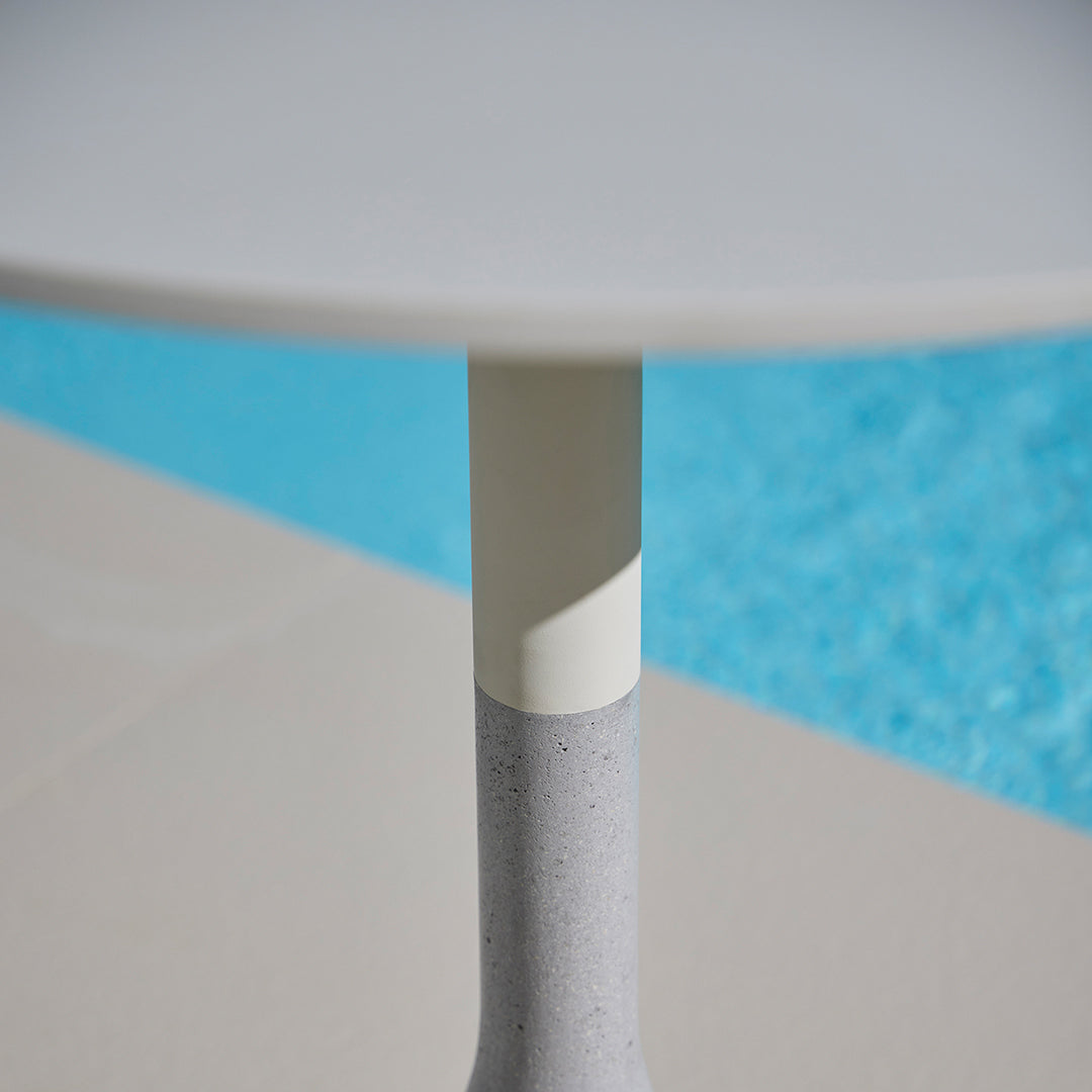 Round table with a smooth white top and textured gray base, set against a vibrant blue pool background.