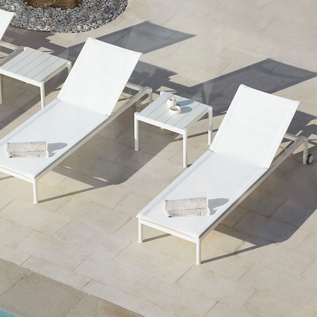 White outdoor lounge chairs and a side table on a light stone patio, showcasing a modern minimalist design.