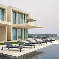 Modern outdoor lounge chairs in sleek gray, set against a light-toned contemporary villa by a blue pool and coastal scenery.
