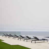 Black lounge chairs lined up on a sunny terrace with green grass and a serene blue ocean backdrop.