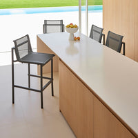 Modern black metal bar stool beside a sleek white countertop, accented by wood cabinetry and green outdoor views.