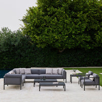 Modern outdoor seating set in gray tones with plush cushions and a sleek black table, surrounded by lush green foliage.