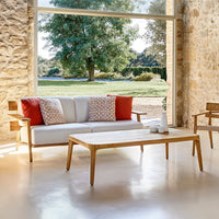 "Modern white sofa with red accent pillows and wooden table, set against a stone wall and green outdoor backdrop."
