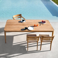 Natural wood table with smooth finish, paired with woven chairs, set against a bright blue pool backdrop.