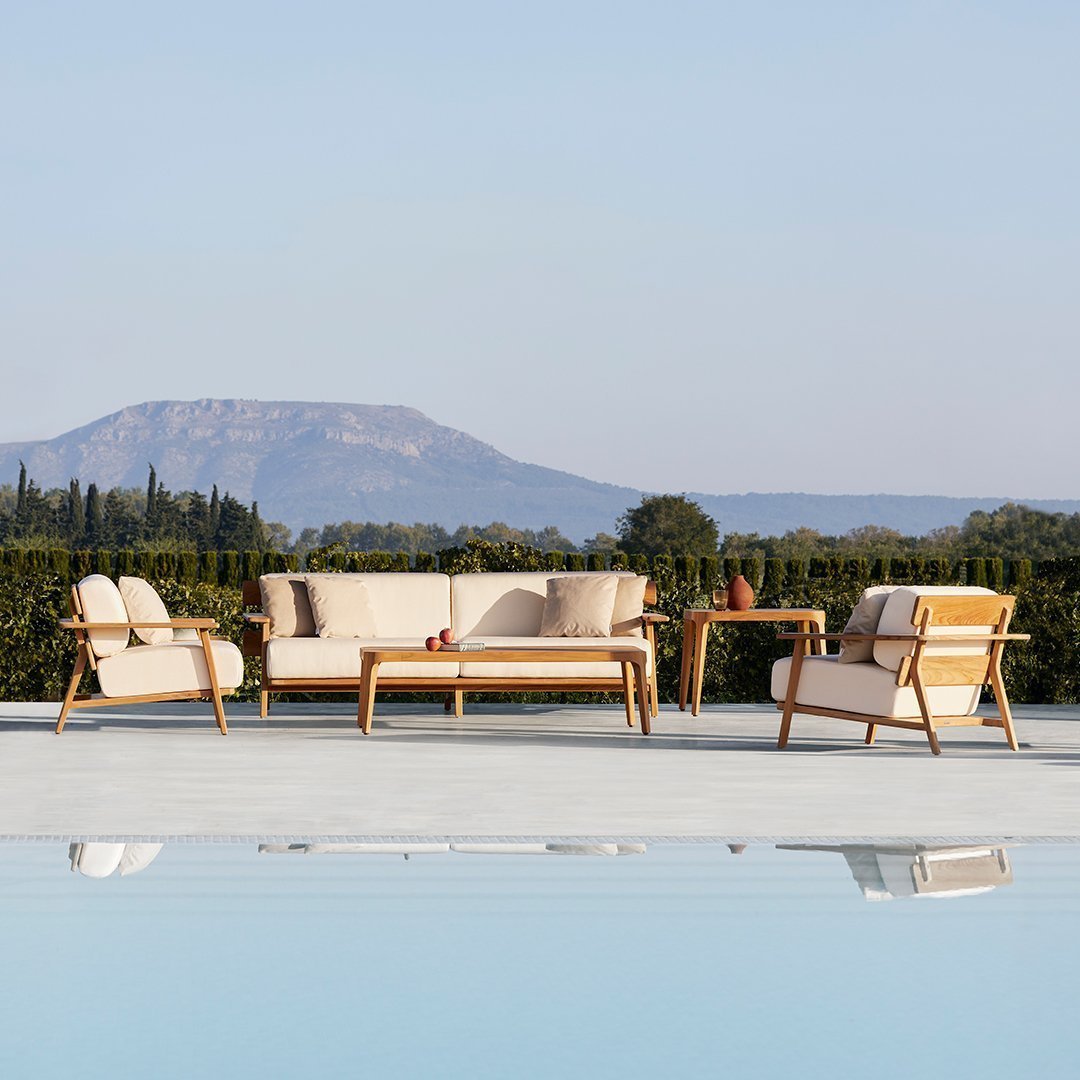 Beige outdoor sofa set with cream cushions and wooden frames, surrounded by lush greenery and a serene pool reflection.