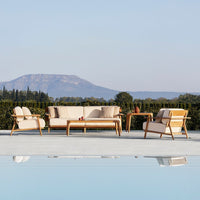 Beige outdoor sofa set with cream cushions and wooden frames, surrounded by lush greenery and a serene pool reflection.