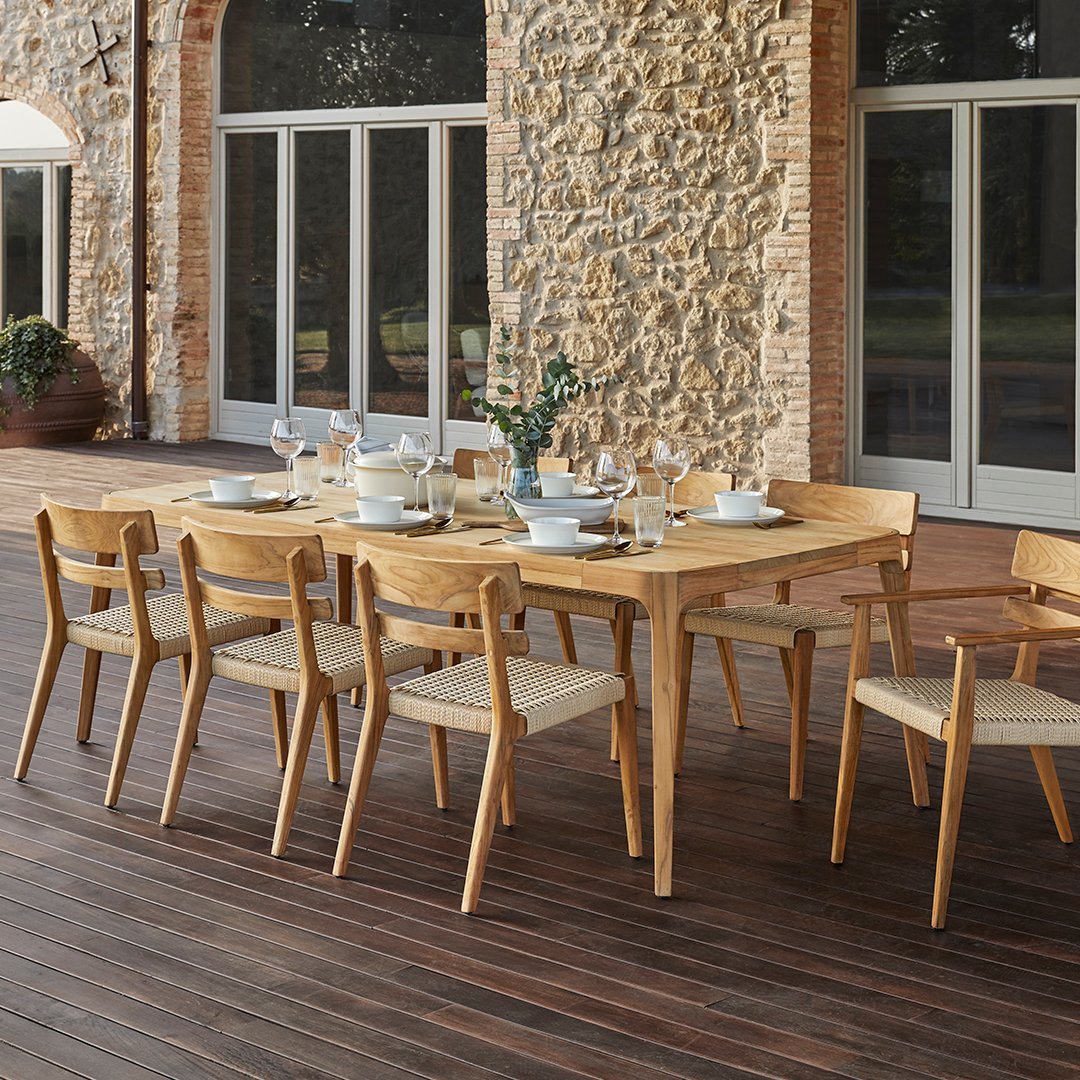 Wooden dining table with natural finish, complemented by beige woven chairs, set against a stone wall backdrop.